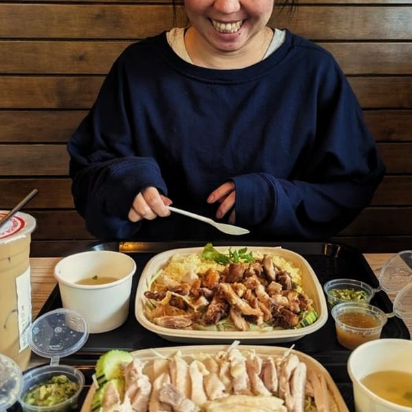 Smiling person holding a spoon, with two trays of Burd Chicken Rice, soup, and drinks.