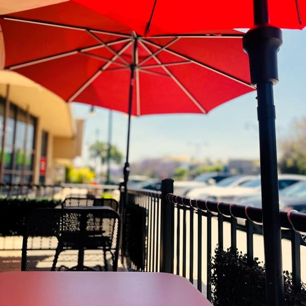 Outdoor patio seating with red umbrellas, black tables, and chairs, overlooking a parking lot.