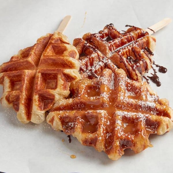 Two golden-brown croffles on sticks, one drizzled with caramel, the other with chocolate and powdered sugar.