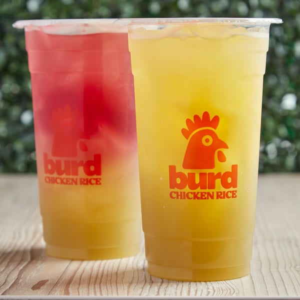 Two Burd Chicken Rice drinks, one red and one yellow, on a wooden table with greenery.