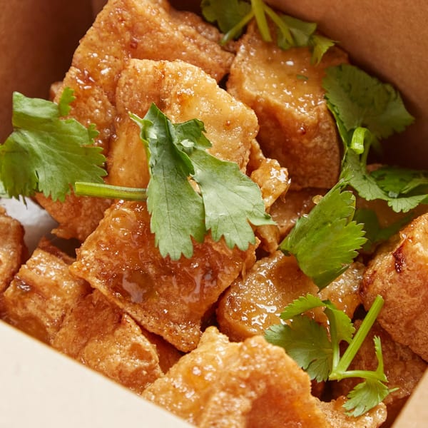 Close-up of crispy fried tofu pieces glazed with sauce and garnished with fresh cilantro in a takeout box.