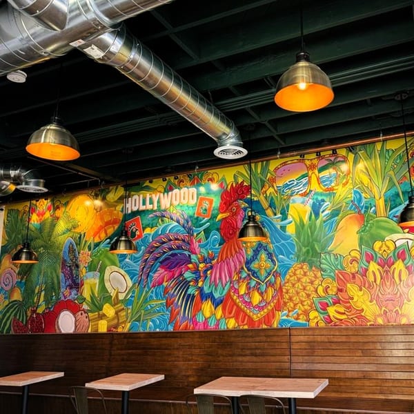 Colorful mural with a rooster, tropical fruits, and "Hollywood" sign above wooden booths and tables.