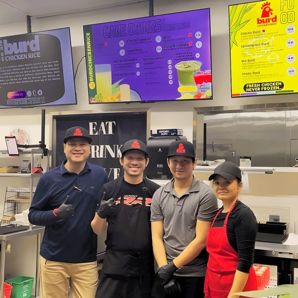 Four Burd Chicken Rice staff members smiling in their kitchen with menu screens behind them.
